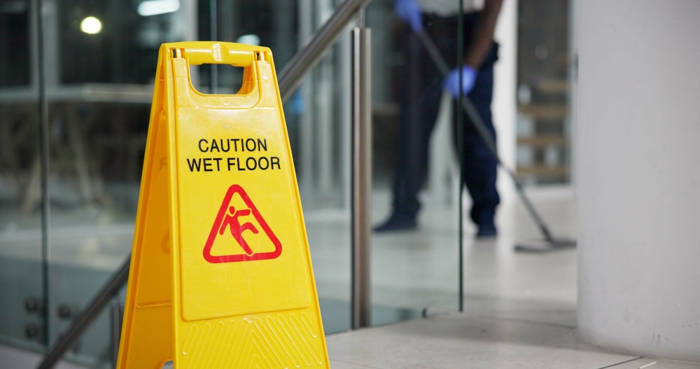 Yellow caution wet floor sign with a cleaner mopping a tiled office entry behind