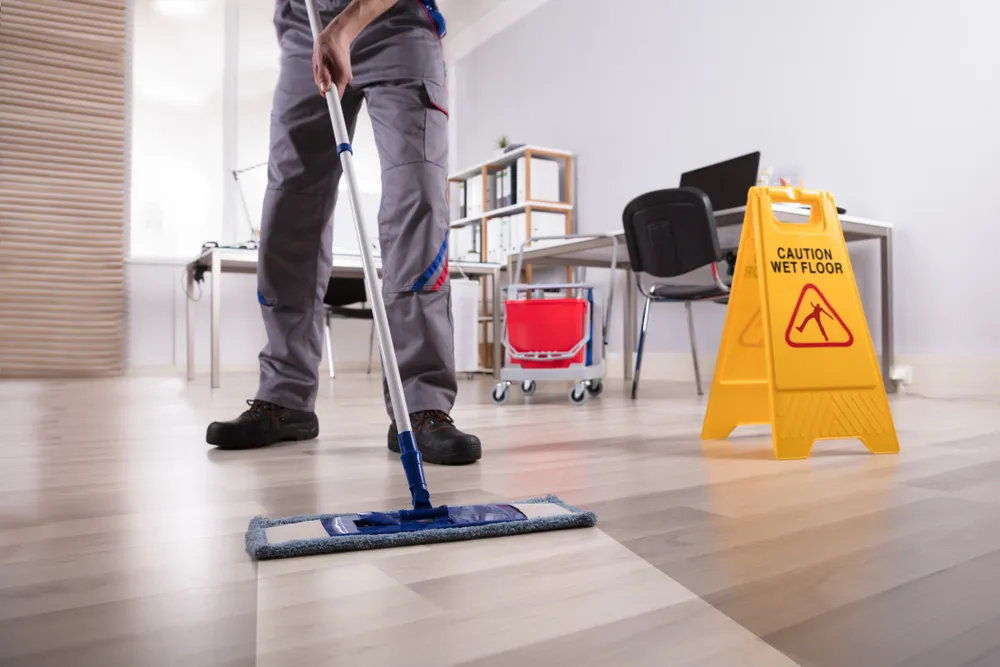 Worker mopping an office floor beside a yellow caution wet floor sign
