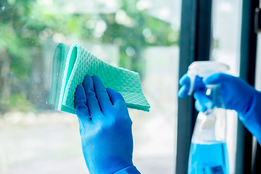 Blue-gloved hand wiping a window with a microfibre cloth and spray bottle