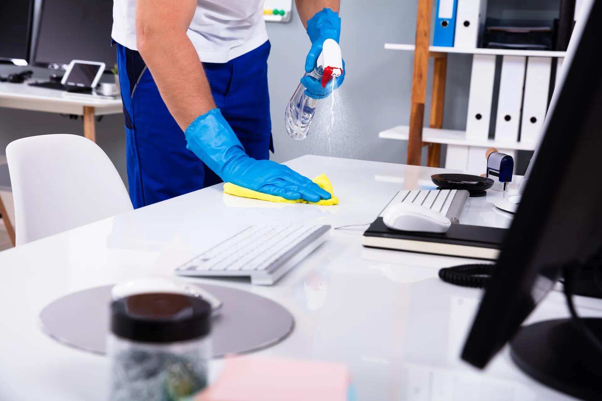 Cleaner in blue gloves spraying and wiping a white office desk with keyboard and monitor in a Townsville workspace